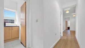 A hallway with white walls, wooden flooring, and a kitchen area with wooden cabinets and a window.