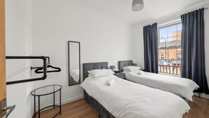A hotel room with two beds, a mirror, a hanging coat rack, a small round table, and a window with curtains.