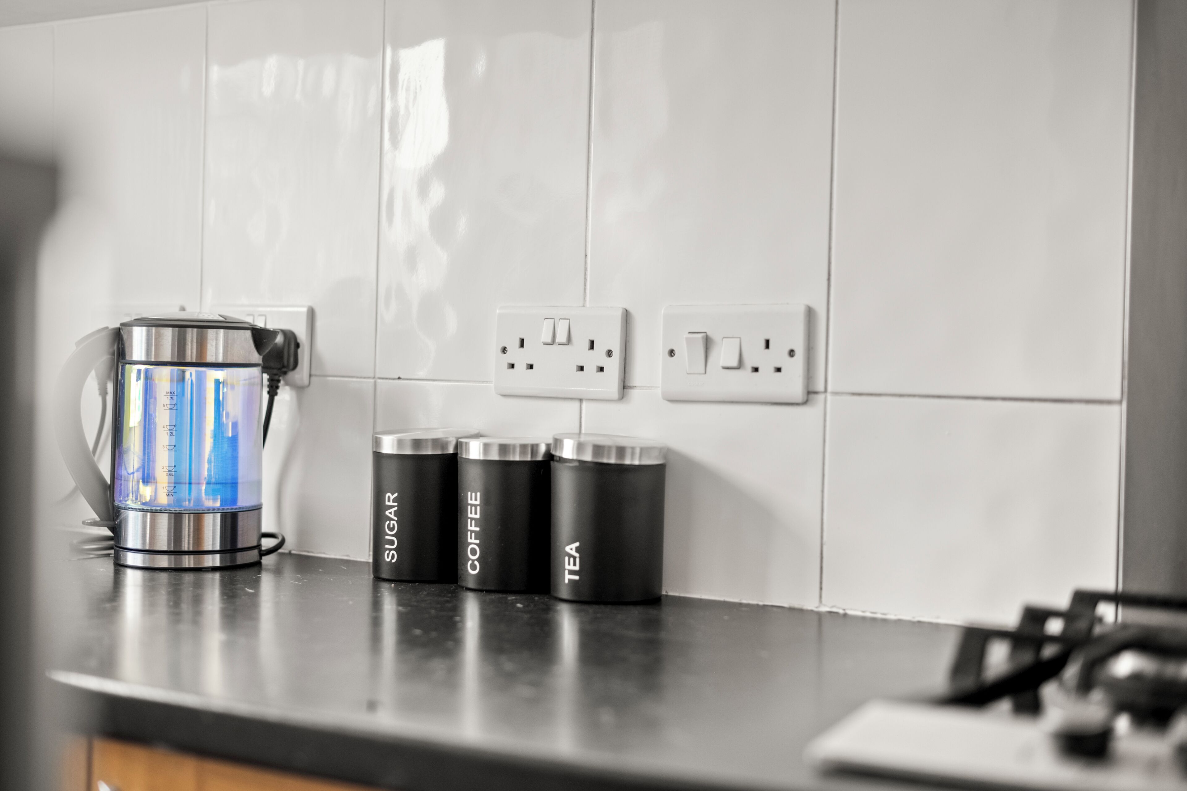 A kitchen with a coffee maker, a kettle, and three containers labeled sugar, coffee, and tea.