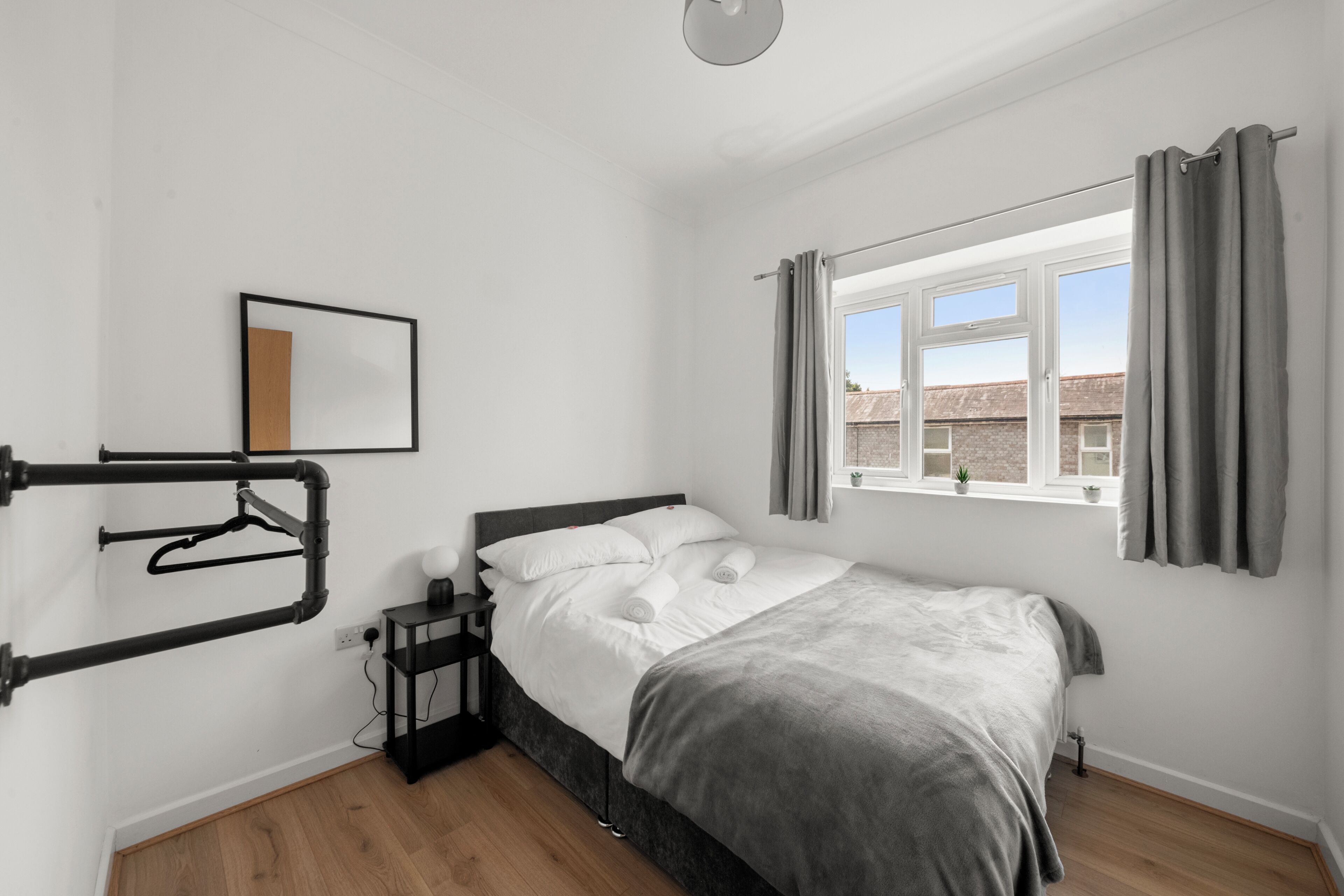 A bedroom with a bed, a nightstand, a window with curtains, and a wall-mounted rack.