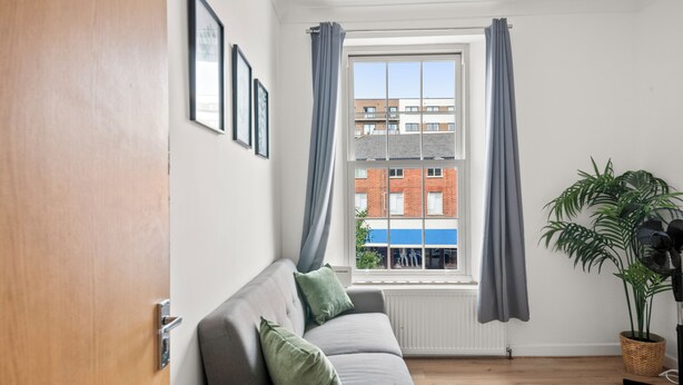 A modern living room with a grey sofa, a large window with curtains, a potted plant, and a radiator.