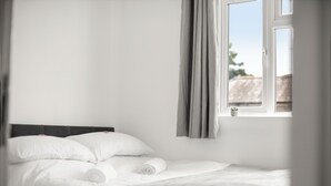 A neatly made bed with white linens and a rolled towel, a window with curtains, and a view of a building outside.