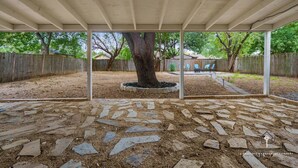 A covered patio area with a stone-tiled floor, a central tree, and a wooden fence in the background. - AT&T, Globe Life, Six Flags - Updated Comfy Home (Grand Prairie)