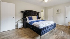 A bedroom with a tufted headboard, a bed with patterned bedding, a white nightstand, a small table with a plant, and a ceiling fan. - AT&T, Globe Life, Six Flags - Updated Comfy Home (Grand Prairie)