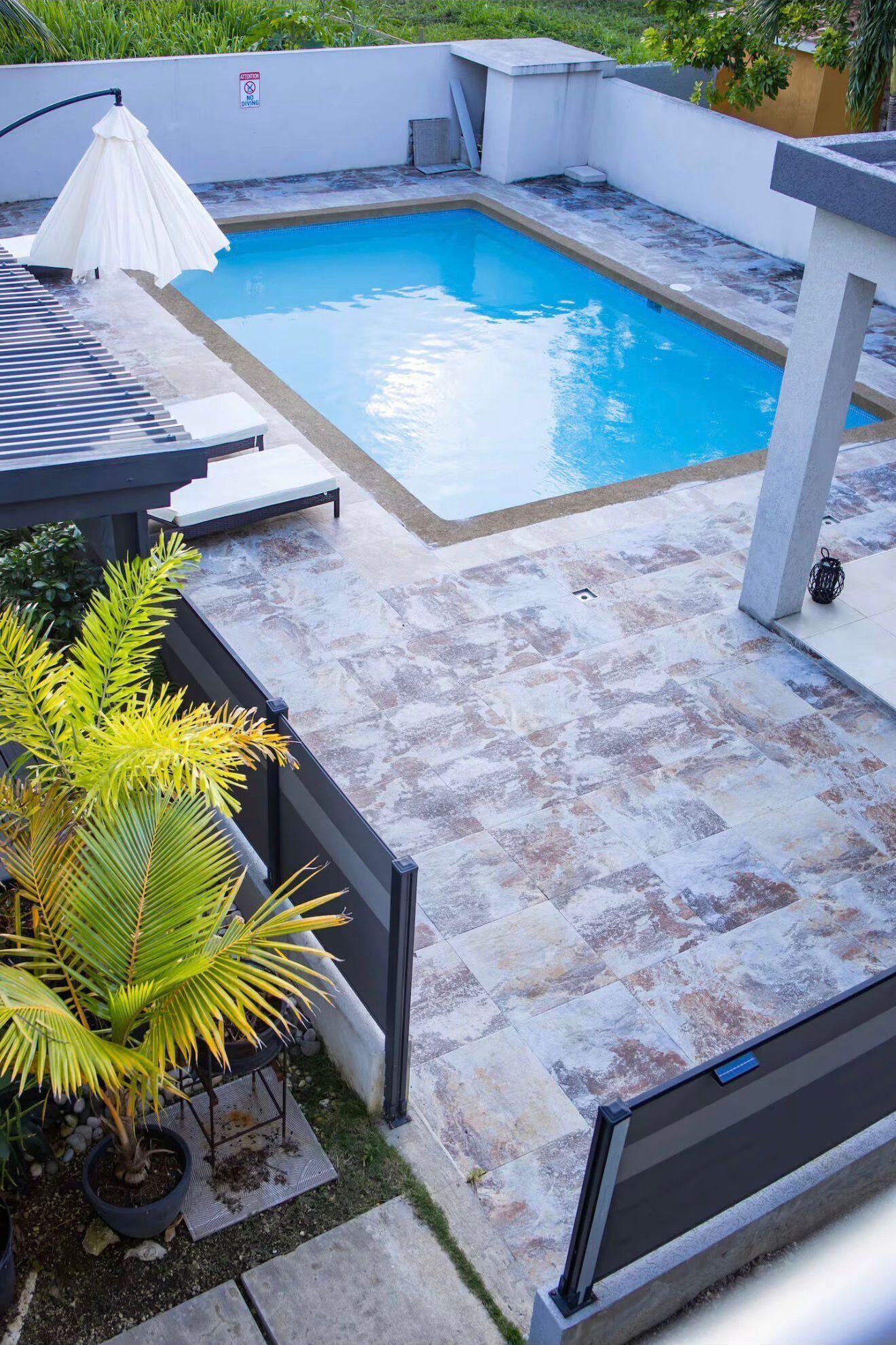 Une piscine rectangulaire, bordée d’une terrasse carrelée, entourée d’un petit muret blanc et agrémentée de quelques plantes en pot.