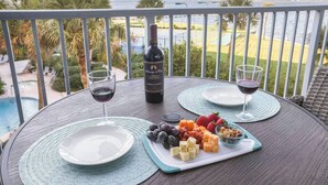 Une table dressée avec une bouteille de vin, deux verres et une planche de fromages agrémentée de fruits et de noix, le tout placé sur un balcon donnant sur une piscine et une végétation luxuriante.
