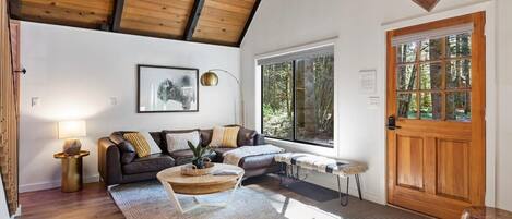 A cozy living room with a wooden ceiling, a large window, a wooden door, and a framed picture on the wall.