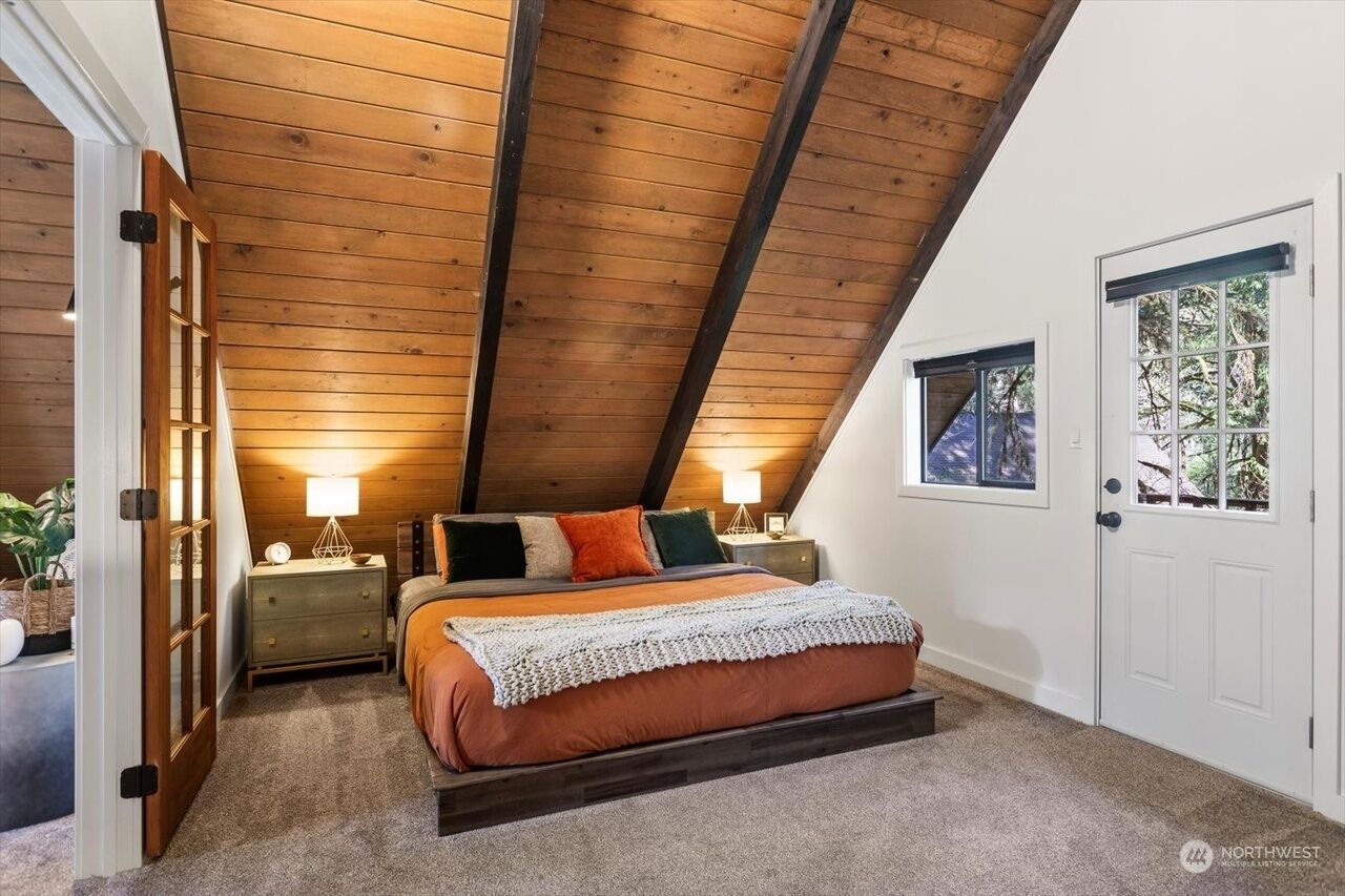A bedroom with a sloped ceiling, a bed with multiple pillows, bedside lamps, and a window with a view of trees.