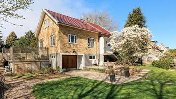 Une maison en pierre à deux étages, avec un toit recouvert de tuiles rouges, un garage à l’avant et un jardin bien entretenu, comprenant une pelouse et un arbre en fleurs.