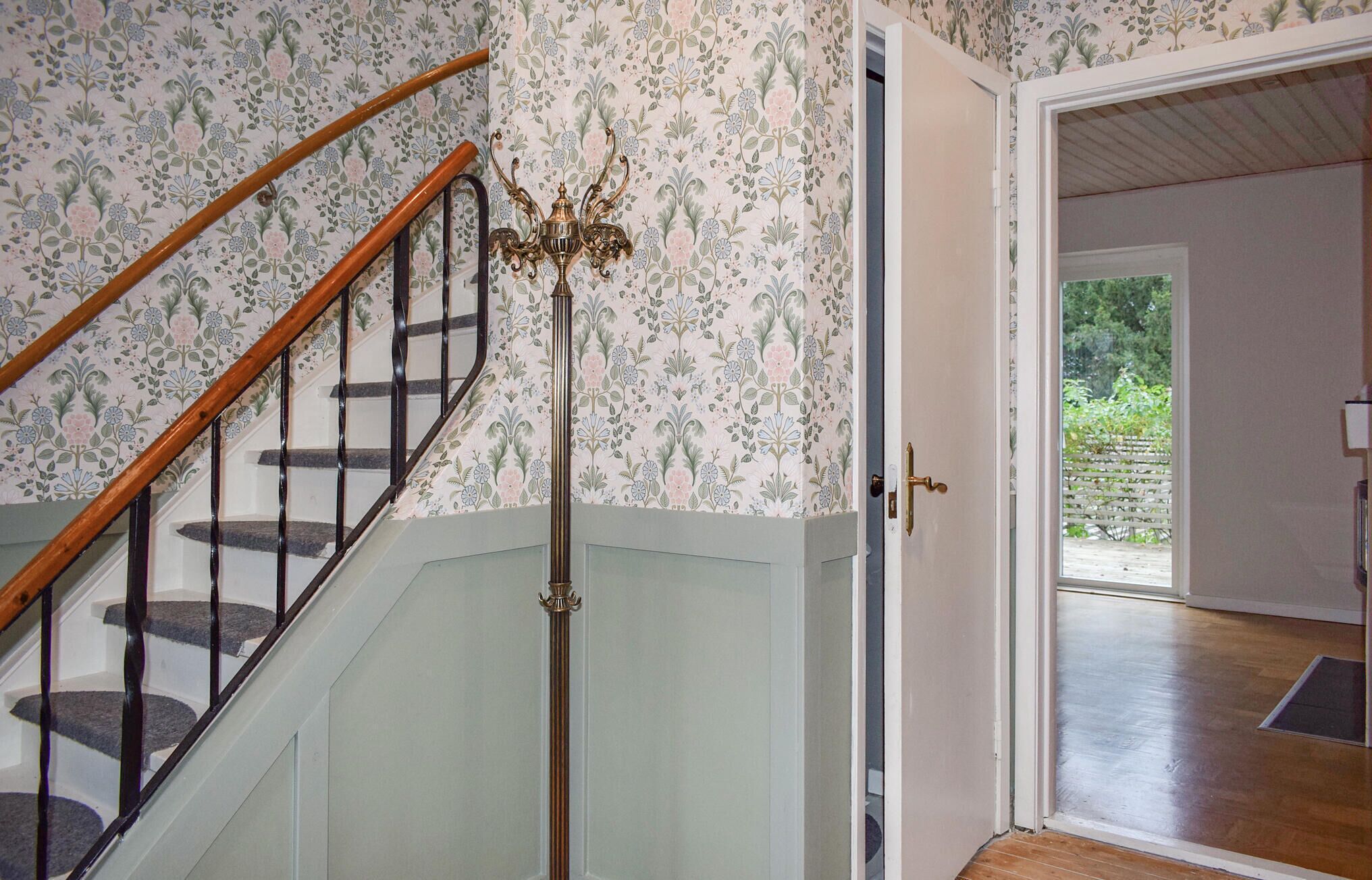 A staircase with a wooden handrail and black balusters, leading to a room with floral wallpaper and a door leading outside.
