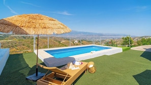 A swimming pool with a view of the landscape, a thatched umbrella, and a wicker lounge chair.