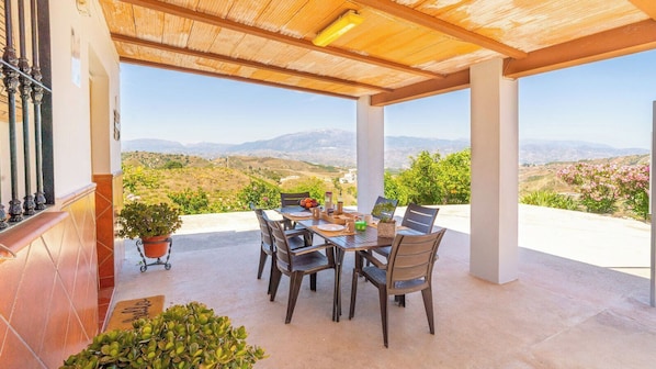 A covered patio with a table and chairs, a potted plant, and a view of the countryside.