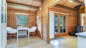 Interior acogedor de una cabaña con detalles de madera, un comedor con dos sillas blancas, una mesa y un ventanal grande.