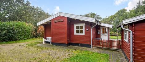 A red wooden building with a white door and window, a bench outside, and a paved area in front.