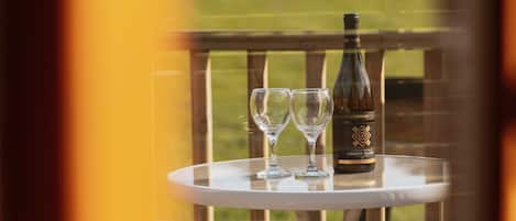 A bottle of wine and two glasses on a small round table with a view of a grassy field through a window.