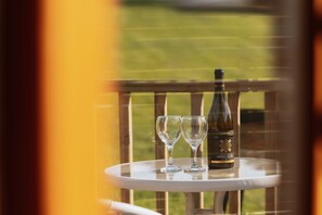 Une bouteille de vin et deux verres sont posés sur une petite table ronde, devant une fenêtre qui donne sur un champ verdoyant.