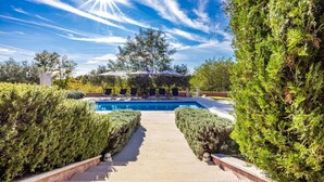 Zona de piscina con cielo despejado, rodeada de setos cuidados y un sendero que lleva a un área de descanso con sombrillas.