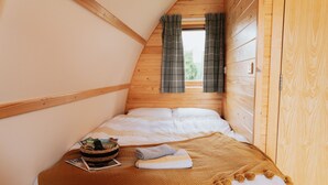 Une chambre confortable avec un plafond en pente en bois, un lit avec une literie blanche, un plaid marron et une petite table sur laquelle se trouvent un panier et un magazine.