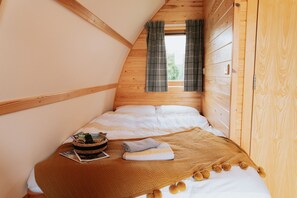 Un dormitorio acogedor con techo inclinado de madera, una cama con ropa blanca, un edredón marrón y una mesita con una cesta y una revista.