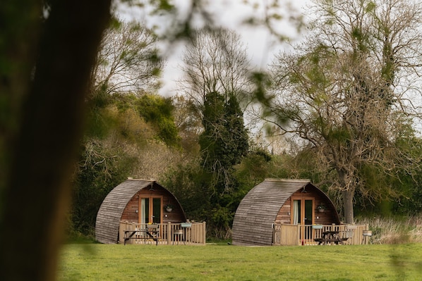 Dos cabañas de madera con forma de cúpula, con terrazas de madera y mesas de picnic, rodeadas de árboles y césped.