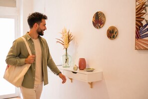 A person walking past a white console table with a vase of dried plants, a red pot, and decorative plates on the wall.