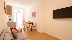 Ein modernes Wohnzimmer mit einer Couch, einem Esstisch mit Stühlen, einem an der Wand befestigten Flachbildfernseher und einer durchgehenden Tür einsehbarer Küchenbereich.