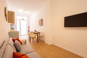 A modern living room with a sofa, a dining table with chairs, a flat-screen TV mounted on the wall, and a kitchen area visible through an open door.