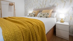 A neatly made bed with a yellow blanket, a bedside table with a lamp, and a wall with a leaf pattern.