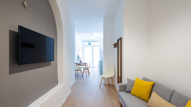 A modern living room with a flat-screen TV mounted on the wall, a grey sofa with yellow cushions, and a wooden dining table with chairs.