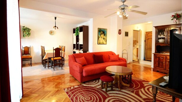 A living room with a red sofa, a dining table with chairs, a ceiling fan, and a kitchen area with wooden cabinets. - Apartment With Scenic View (Porec)