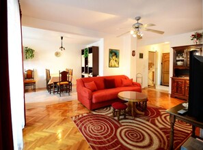 A living room with a red sofa, a dining table with chairs, a ceiling fan, and a kitchen area with wooden cabinets. - Apartment With Scenic View (Porec)