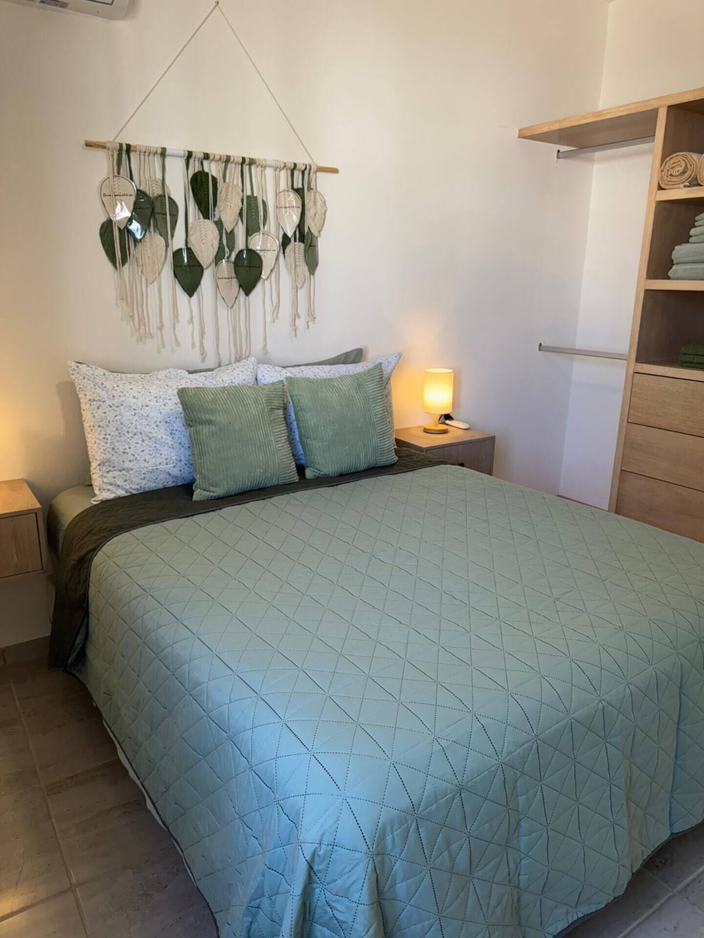 Ein ordentlich bezogenes Bett mit einer gesteppten, grünen Tagesdecke, einem Wandbehang aus Makramee und einer Nachttischlampe.