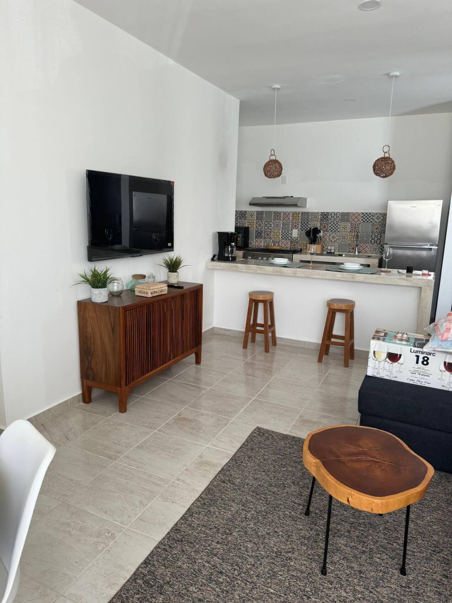 Cocina moderna con televisor de pantalla plana, cafetera y refrigerador. Hay dos taburetes y un armario de madera con plantas.