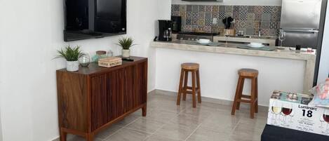 Cocina moderna con televisor de pantalla plana, cafetera y refrigerador. Hay dos taburetes y un armario de madera con plantas.