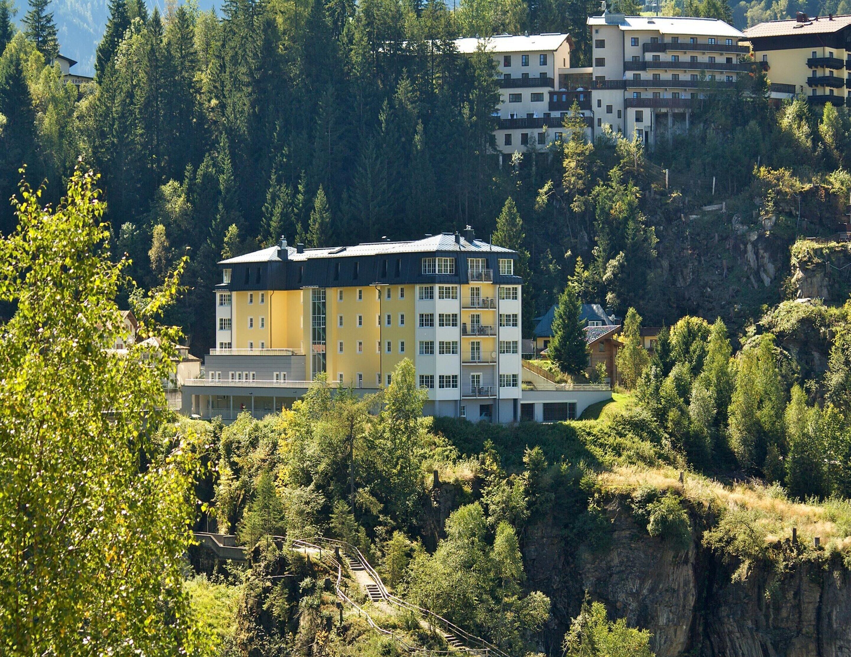 A multi-story hotel with balconies, surrounded by dense forest and a steep cliff.