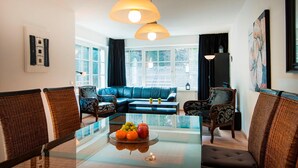Un salón moderno con una mesa de comedor de vidrio, sillas de mimbre, un sofá azul y ventanas grandes con cortinas.
