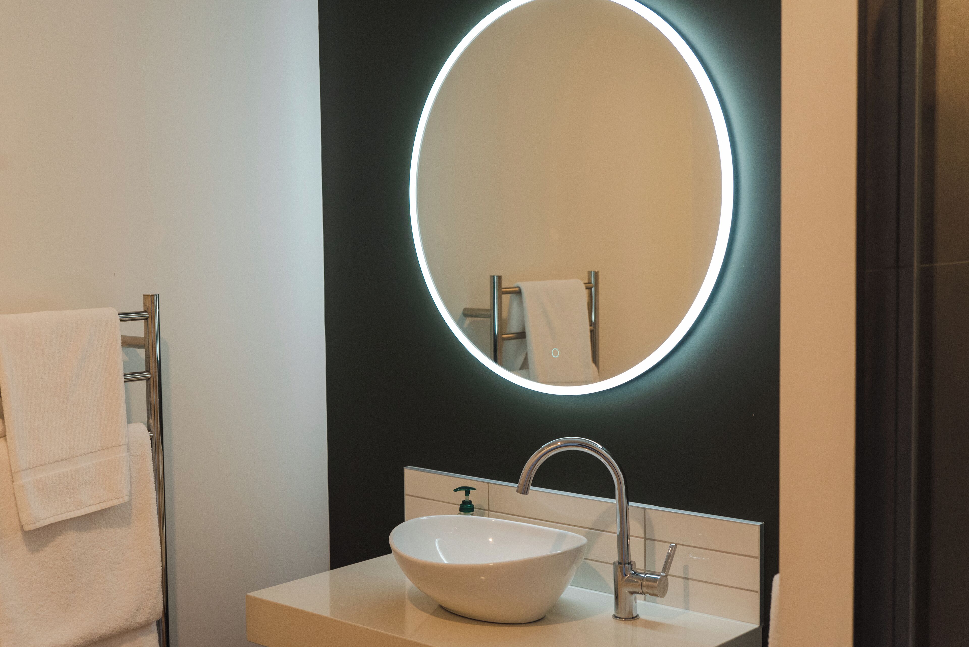 Un baño moderno con un lavamanos blanco, un espejo redondo con luz y un perchero para toallas.