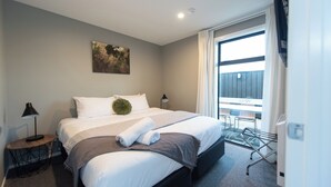Habitación de hotel moderna con una cama grande, una mesita de noche con lámpara, un cuadro en la pared y vistas al balcón exterior.