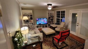 Una sala de estar con televisor, sofá, reposapiés y una silla roja. Se observan estanterías y una puerta al fondo.