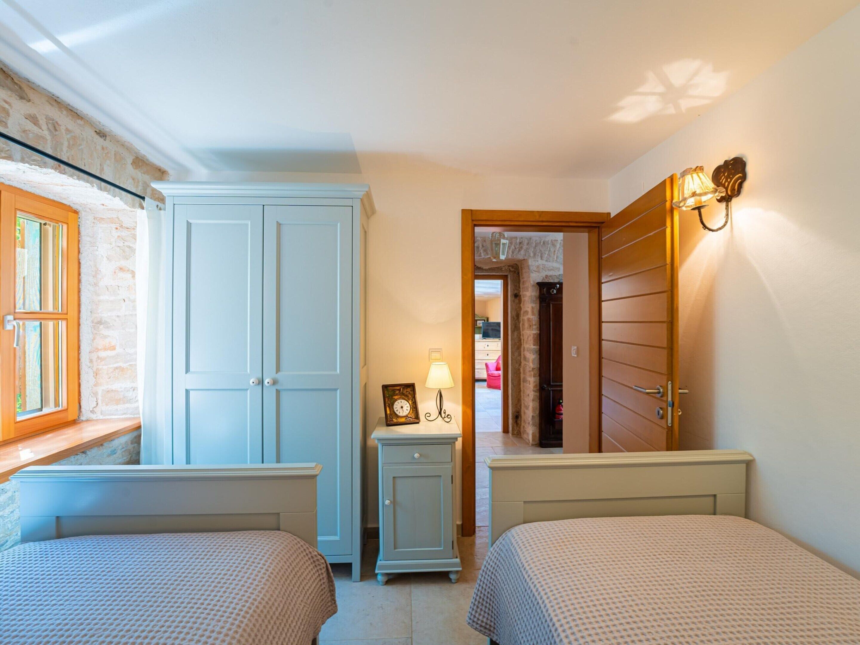 A bedroom with two beds, a wardrobe, a nightstand with a lamp, and a door leading to another room.