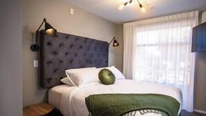 Een hotelkamer met een gestapeld hoofdbord, een bed met witte lakens, een groene plaid, een klein rond tafeltje en een plafondlamp.