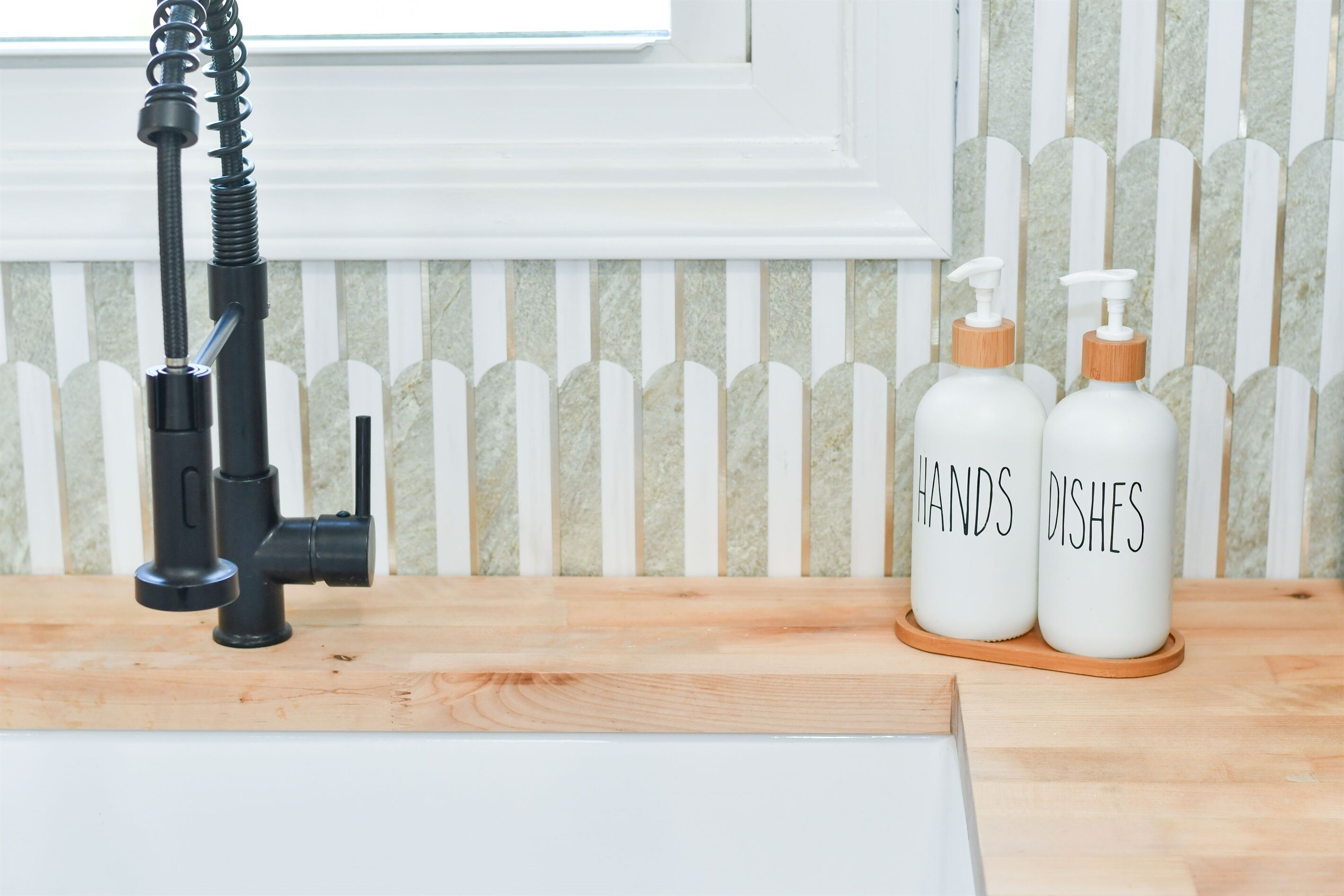 A kitchen with a black faucet, two white bottles labeled "HANDS Dishes" on a wooden tray, and a white picket fence in the background.