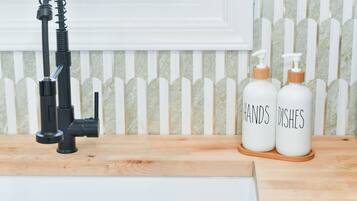 A kitchen with a black faucet, two white bottles labeled "HANDS Dishes" on a wooden tray, and a white picket fence in the background.