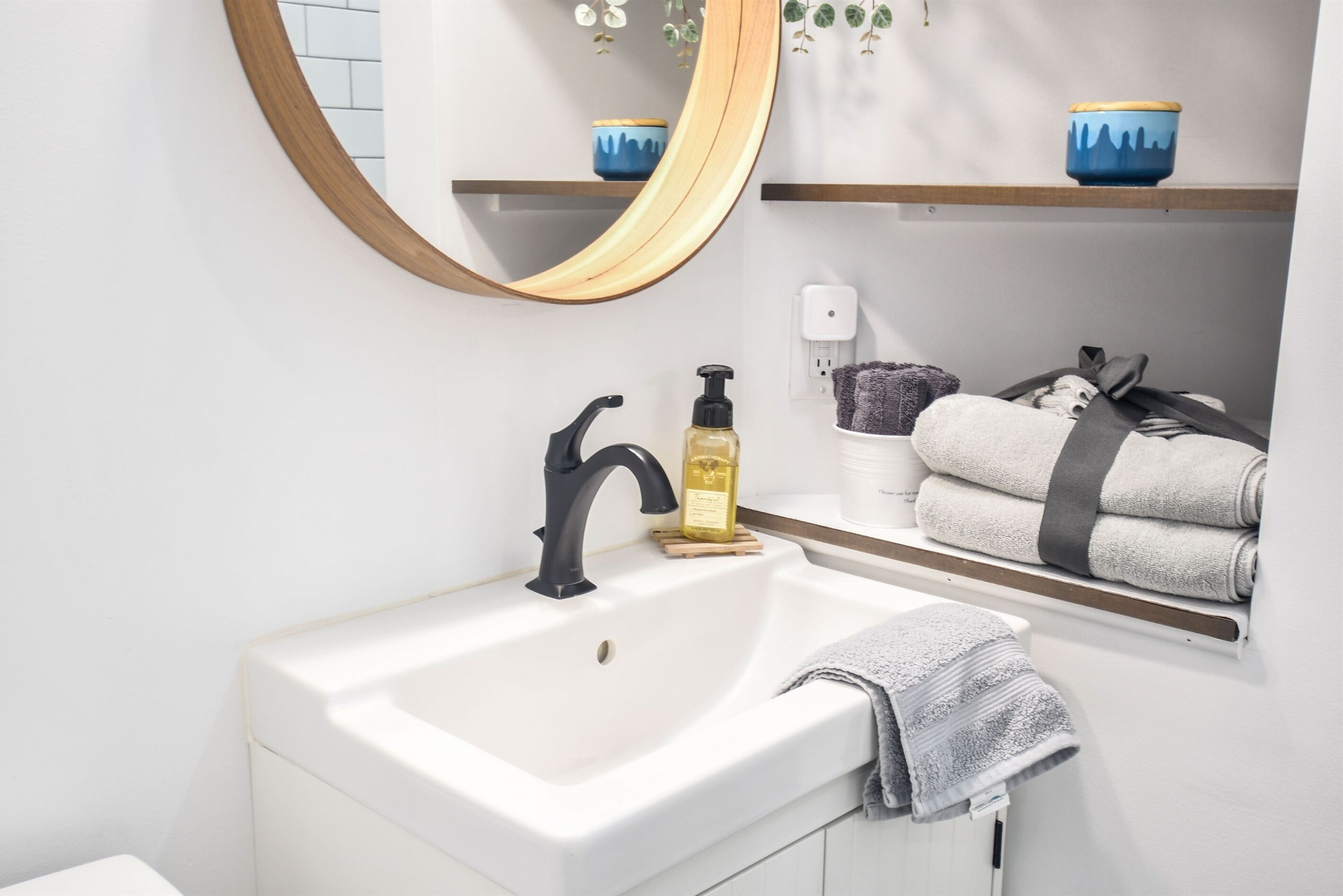 Ein Badezimmer mit einem weißen Waschbecken, einem schwarzen Wasserhahn und einem Spiegel mit Holzrahmen. Handtücher und ein Seifenspender befinden sich auf Regalen.