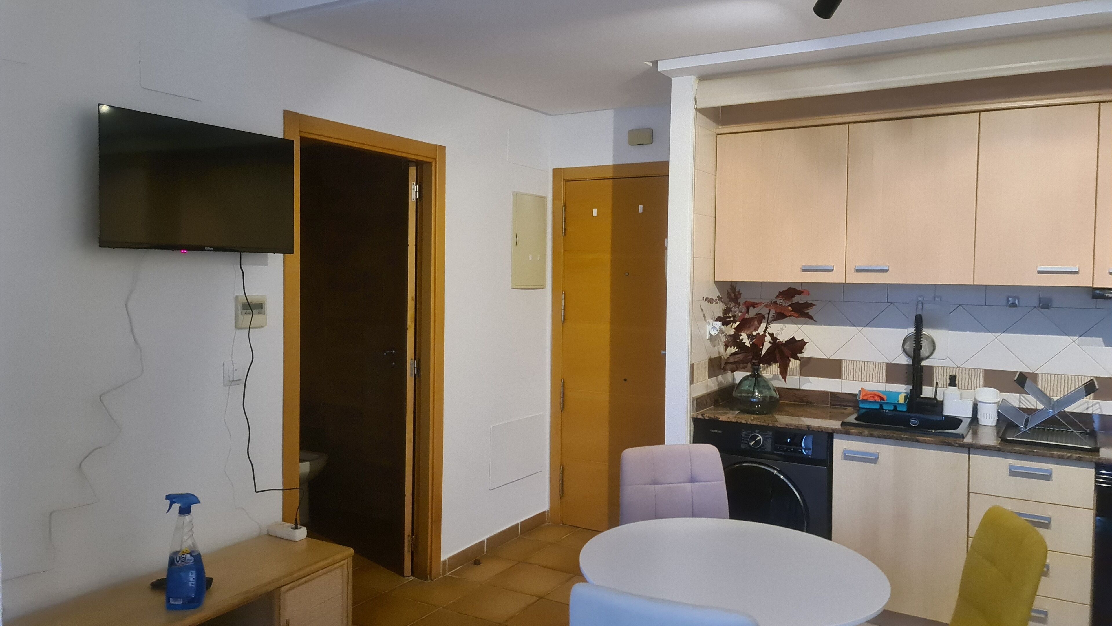 A compact kitchen with wooden cabinets, a built-in dishwasher, and a small dining table with a chair.