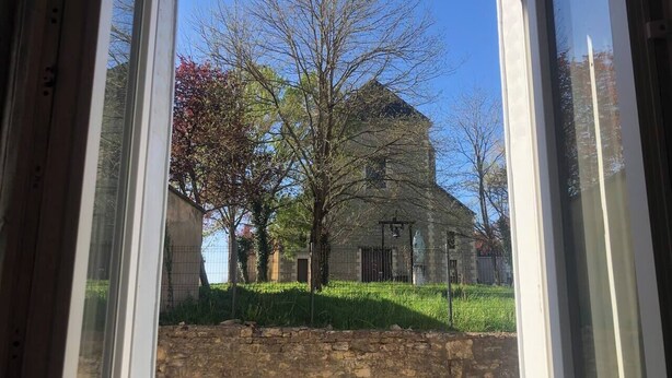 A view through a window of a stone building with a grassy area and trees in front. - 3 rooms flat with garden next to the train station (Nevers)