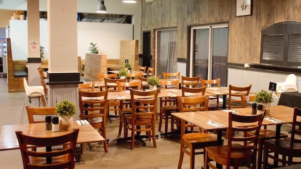 A restaurant with wooden tables and chairs, potted plants, and a view of the kitchen area. - The Caledonian Hotel (Wonthaggi)