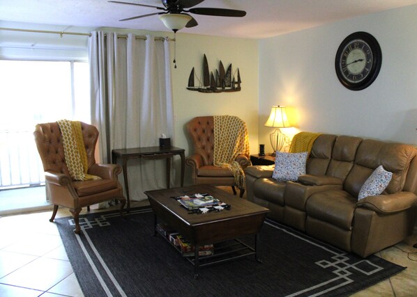 A living room with a brown leather sofa, two armchairs, a coffee table, a lamp, a clock, and a window with curtains.