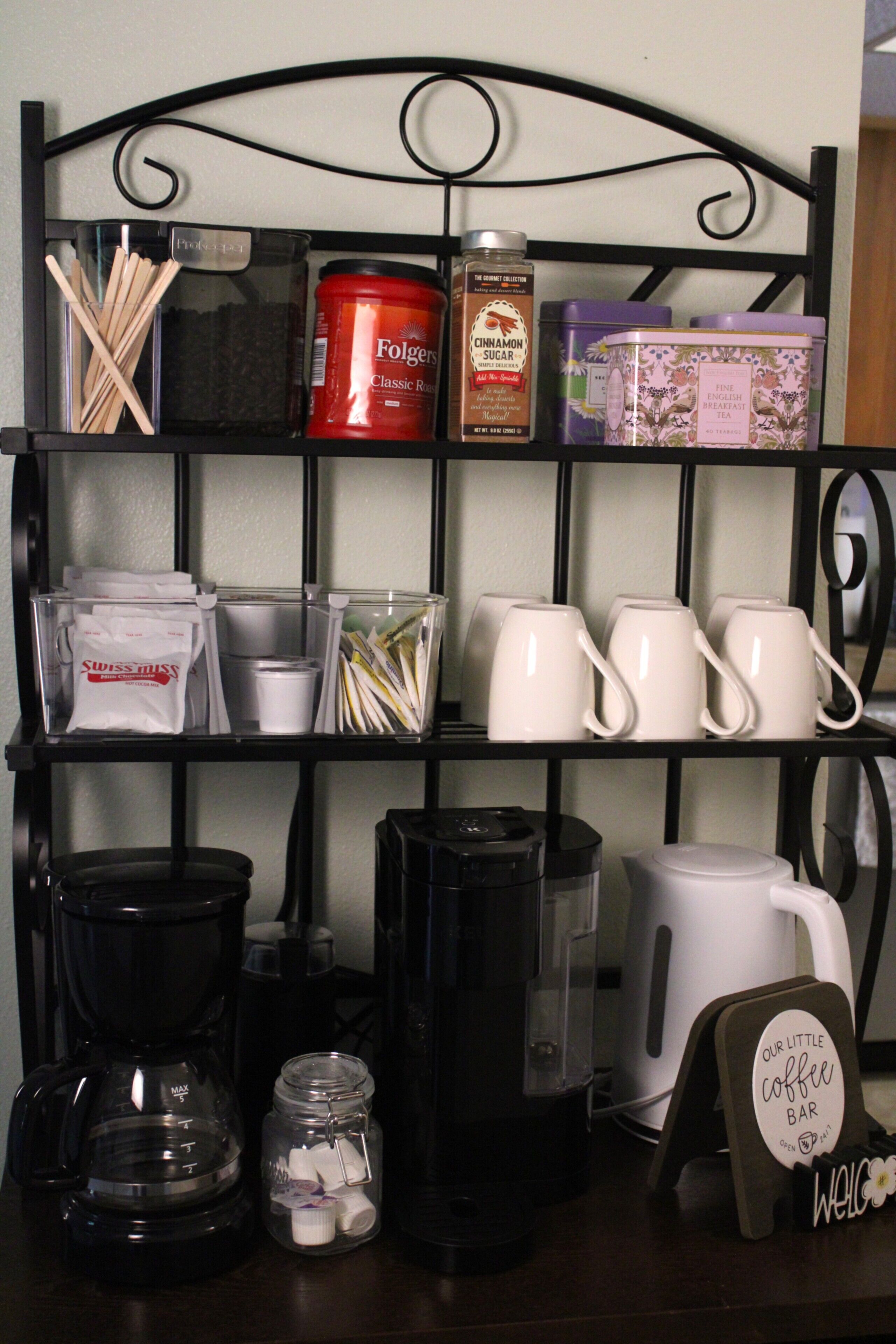 A coffee station with a coffee maker, cups, and a sign that reads 'Our Little Coffee Bar.'