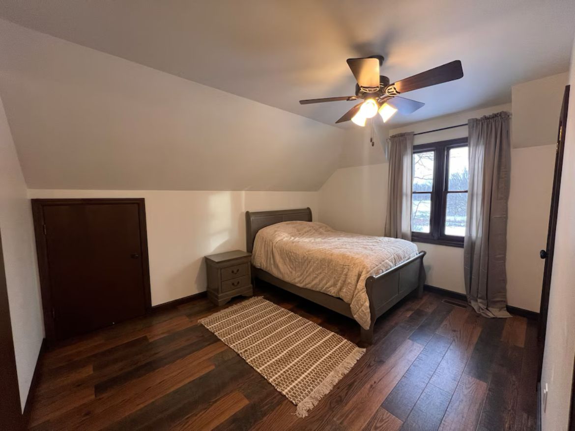 A bedroom with a bed, a ceiling fan, a window with curtains, and a wooden floor.